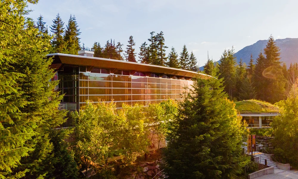 Side view of the Squamish Lil'wat Cultural Centre in summer, featuring a vibrant green roof and forest surroundings.