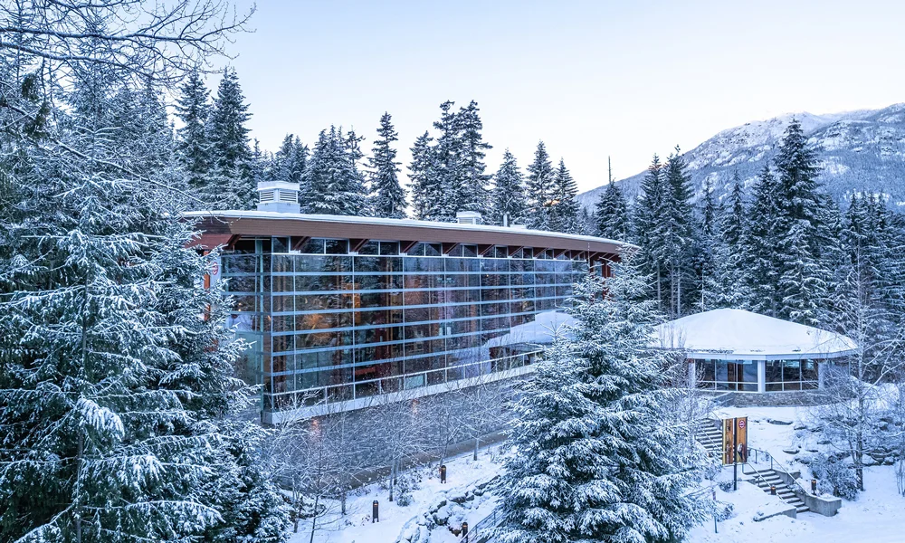 Side view of the Squamish Lil'wat Cultural Centre in winter, with snow-draped trees and expansive glass architecture.